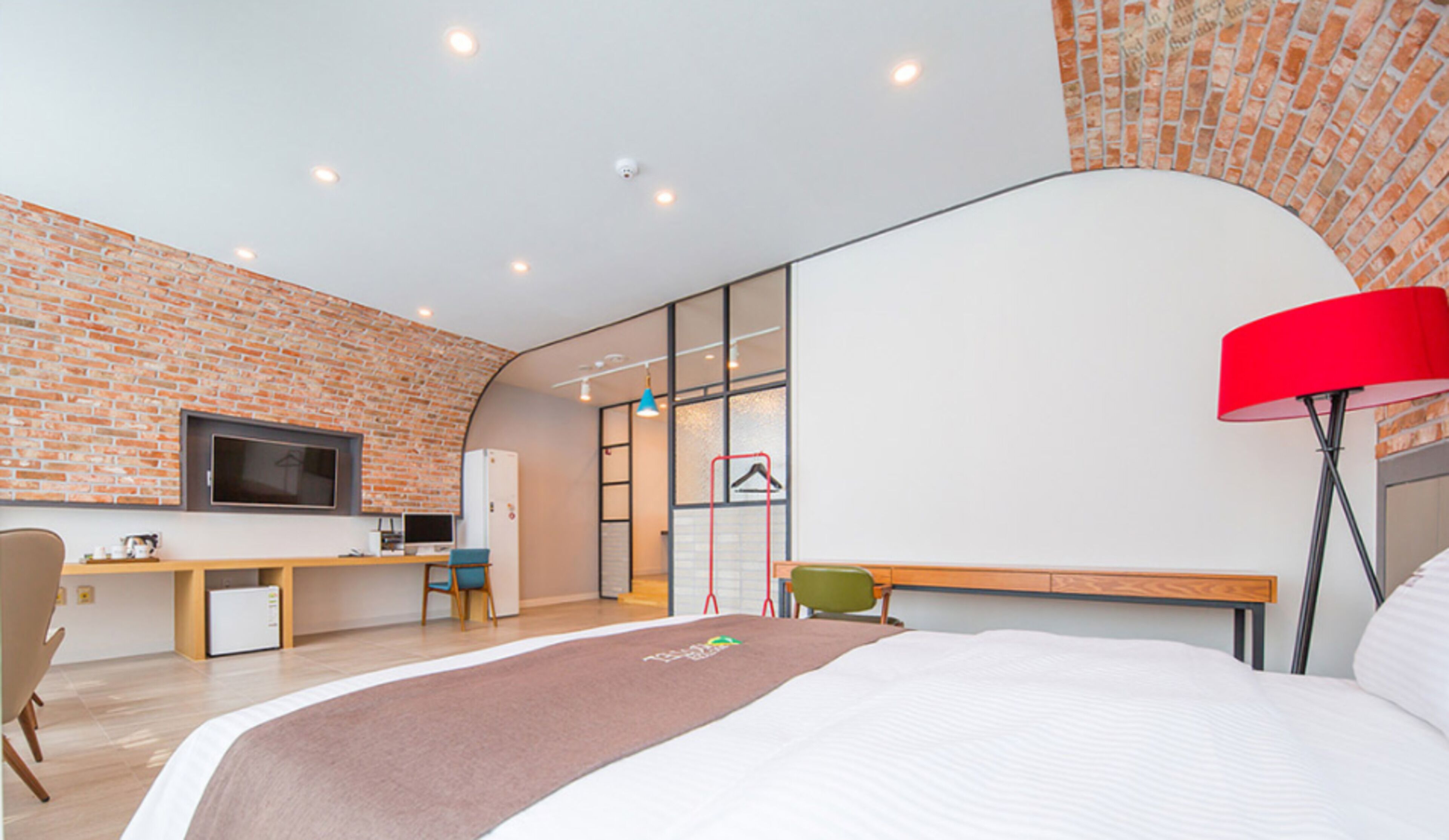 A modern hotel room with a brick wall, a bed, a desk, a chair, a TV, and a red lamp.