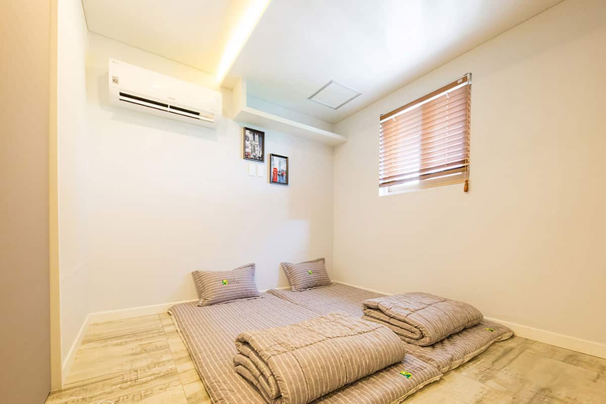 Ein Schlafzimmer mit Bett, Kissen, einem Fenster mit Jalousien, einer Klimaanlage und einem kleinen gerahmten Bild an der Wand.