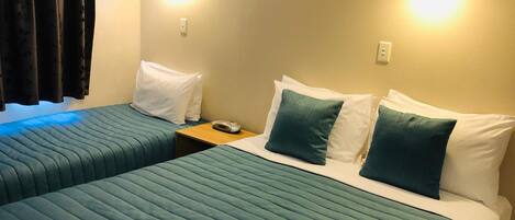 Habitación de hotel con dos camas, cada una con cubrecama color turquesa, almohadas blancas y una mesita de noche con lámpara.