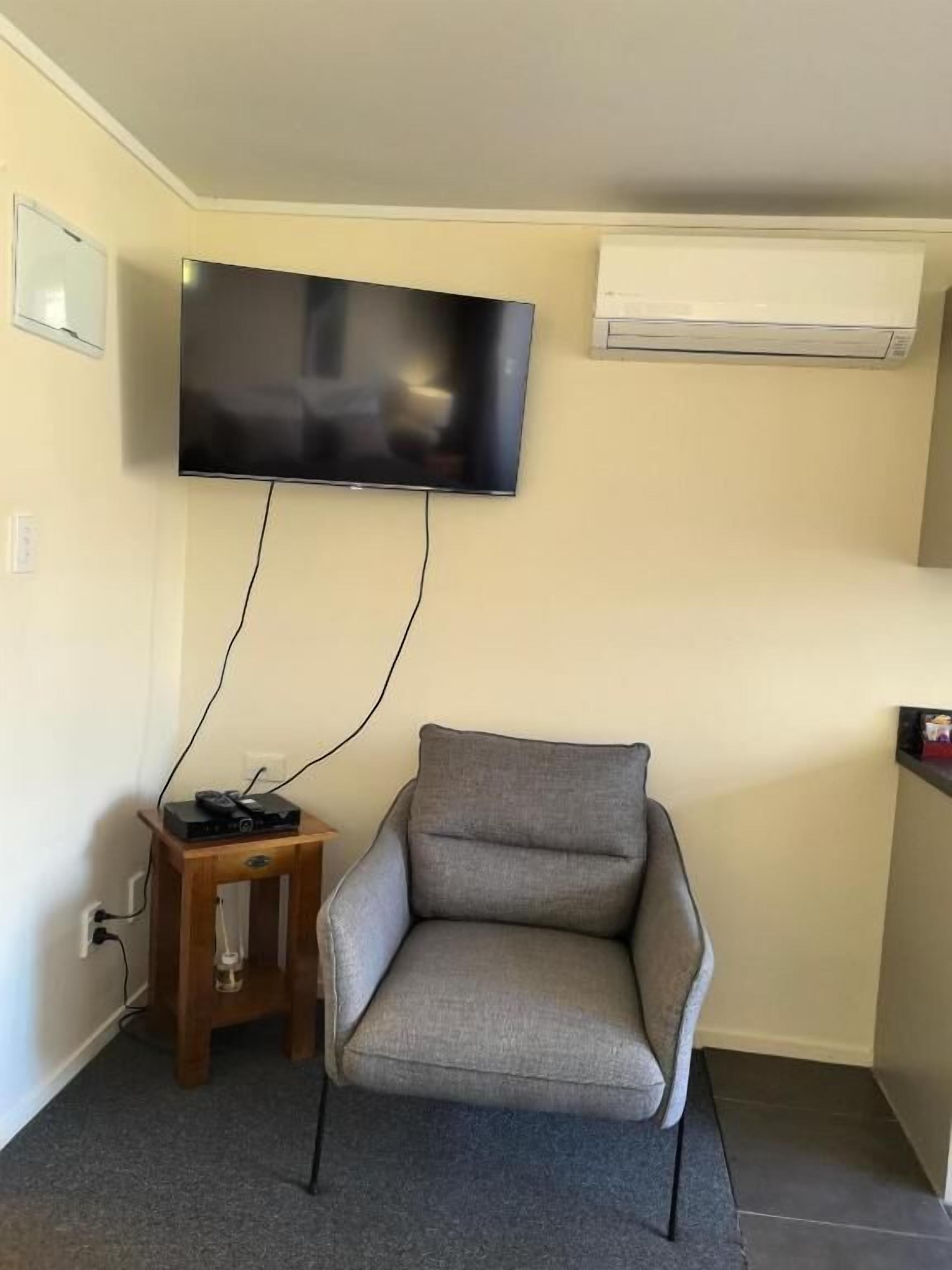 A room with a flat-screen TV mounted on the wall, a grey armchair, a small wooden side table with a glass, and an air conditioning unit.