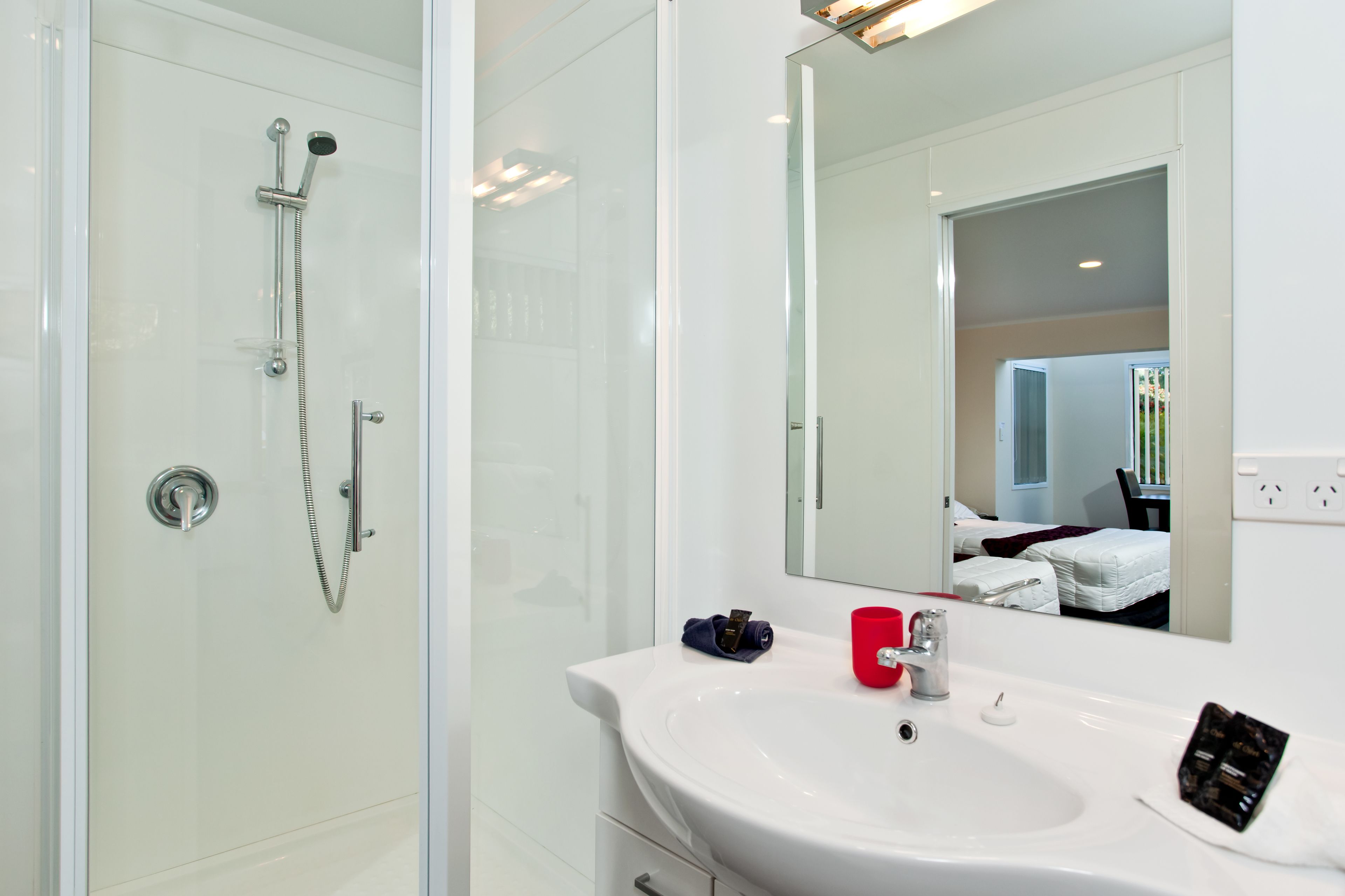 Un baño moderno con cabina de ducha de vidrio, un lavamanos blanco y una taza roja, y un espejo grande que refleja un dormitorio.