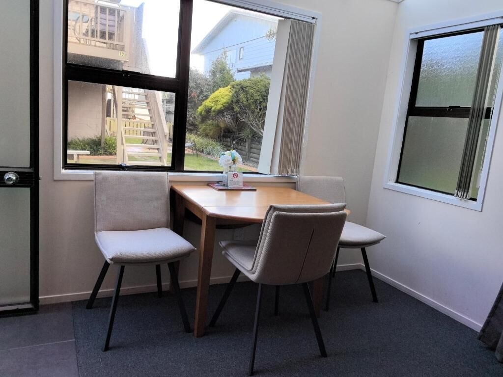 A dining area with a table and four chairs, a window with a view of a residential building, and a door with a handle.