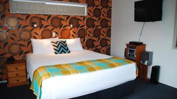 A neatly made bed with a colorful blanket, a wall-mounted TV, and a small speaker on a wooden stand.