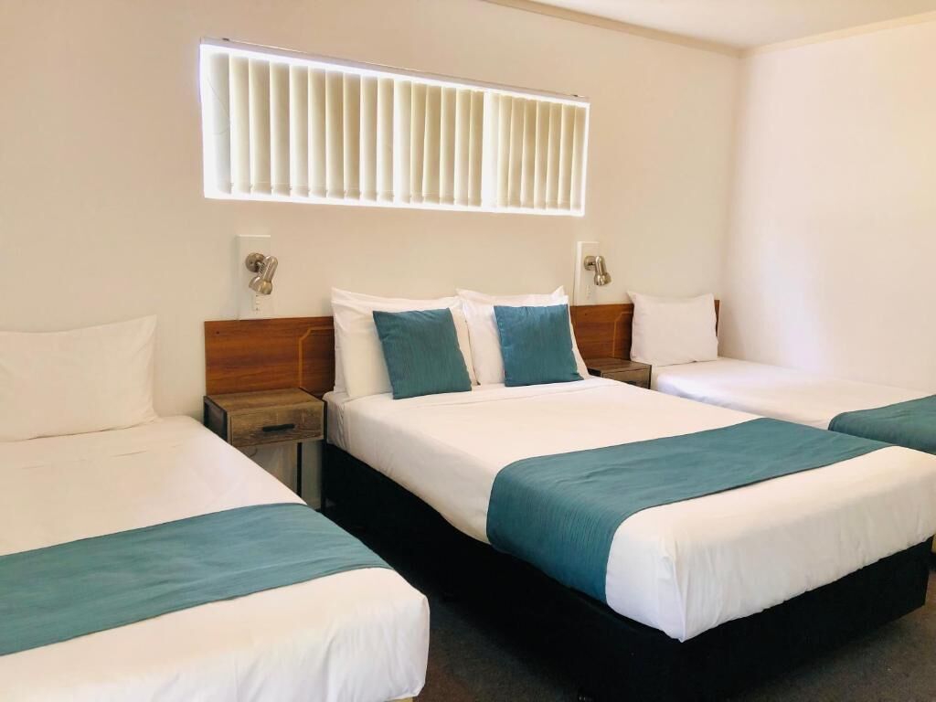 Habitación de hotel con tres camas dispuestas en fila, una ventana con persianas y un aplique de pared.
