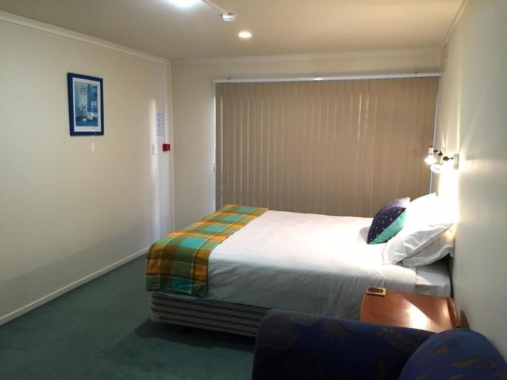 A hotel room with a bed, a chair, a bedside table, a lamp, and a window with blinds.