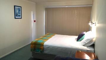A hotel room with a bed, a chair, a bedside table, a lamp, and a window with blinds.