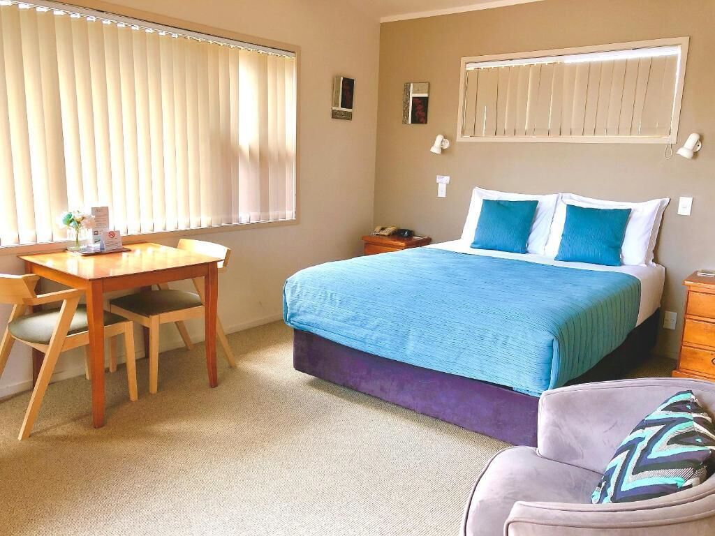 Ein Hotelzimmer mit einem Bett, einem Schreibtisch und einem Stuhl. Es gibt ein Fenster mit Jalousien, einen Nachttisch und eine Lampe.