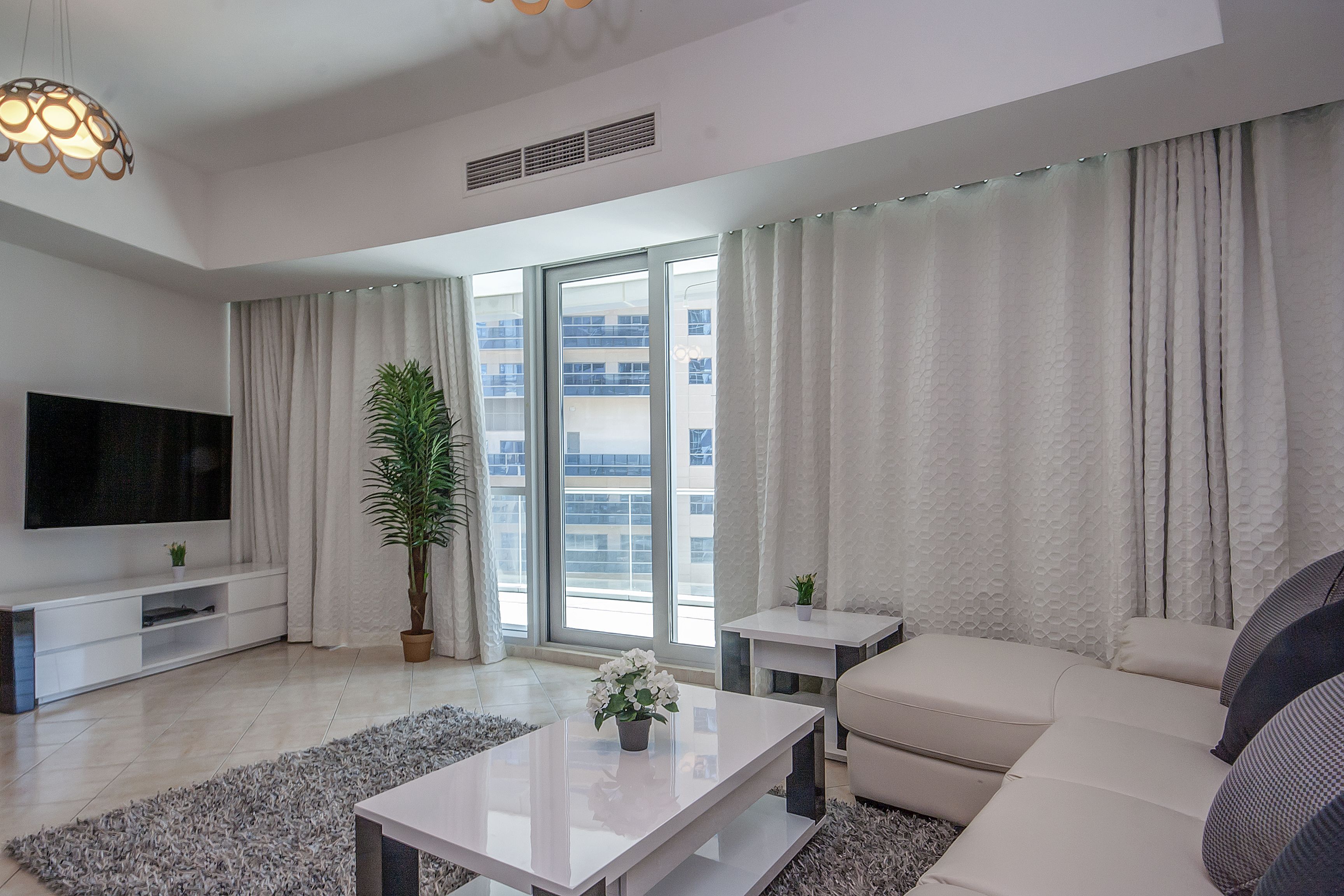 A modern living room with a flat-screen TV, a sofa, a coffee table, a potted plant, and large windows with curtains.