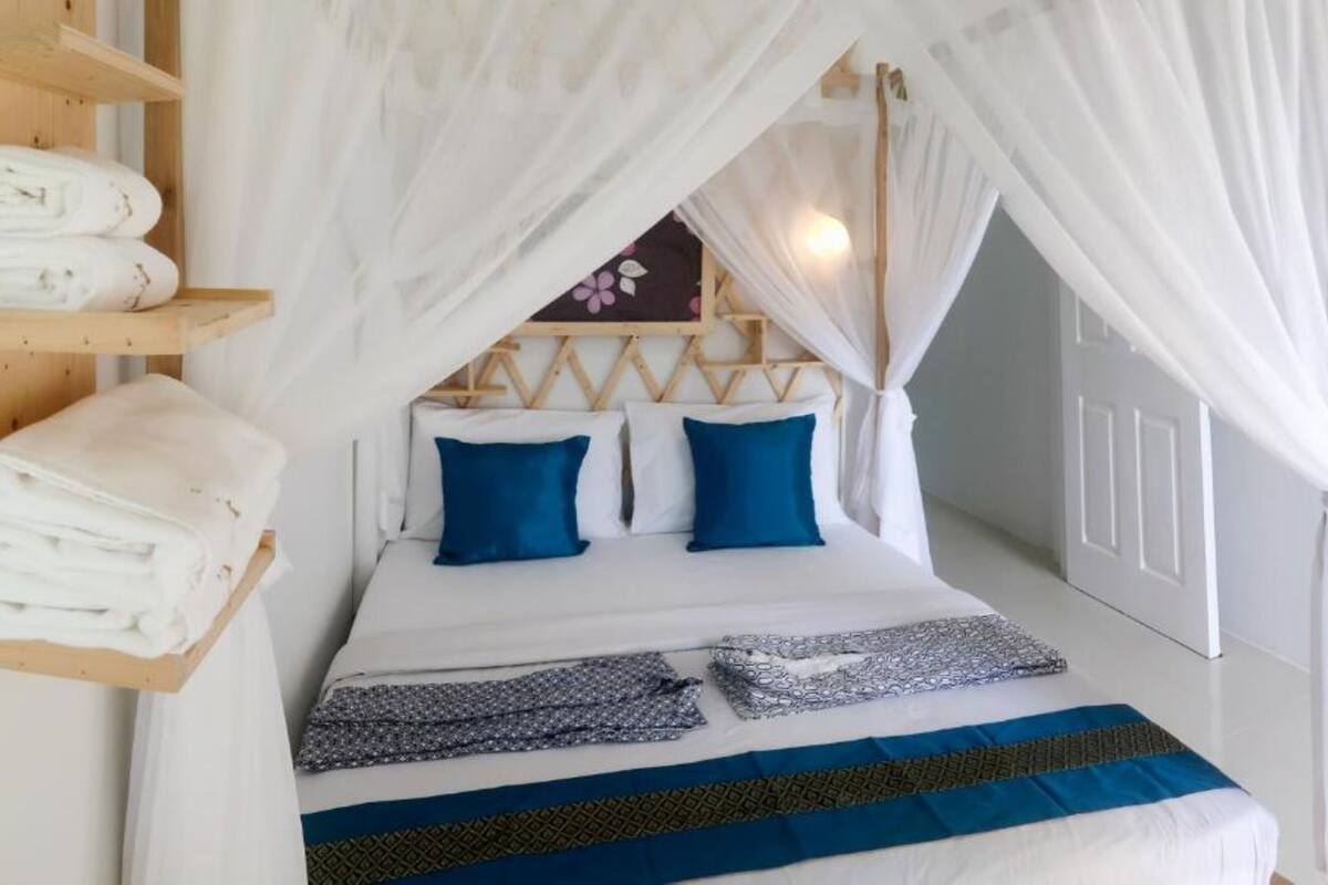 Un lit bien fait, surmonté d’un dais, une étagère avec des serviettes pliées et une couverture à motifs.
