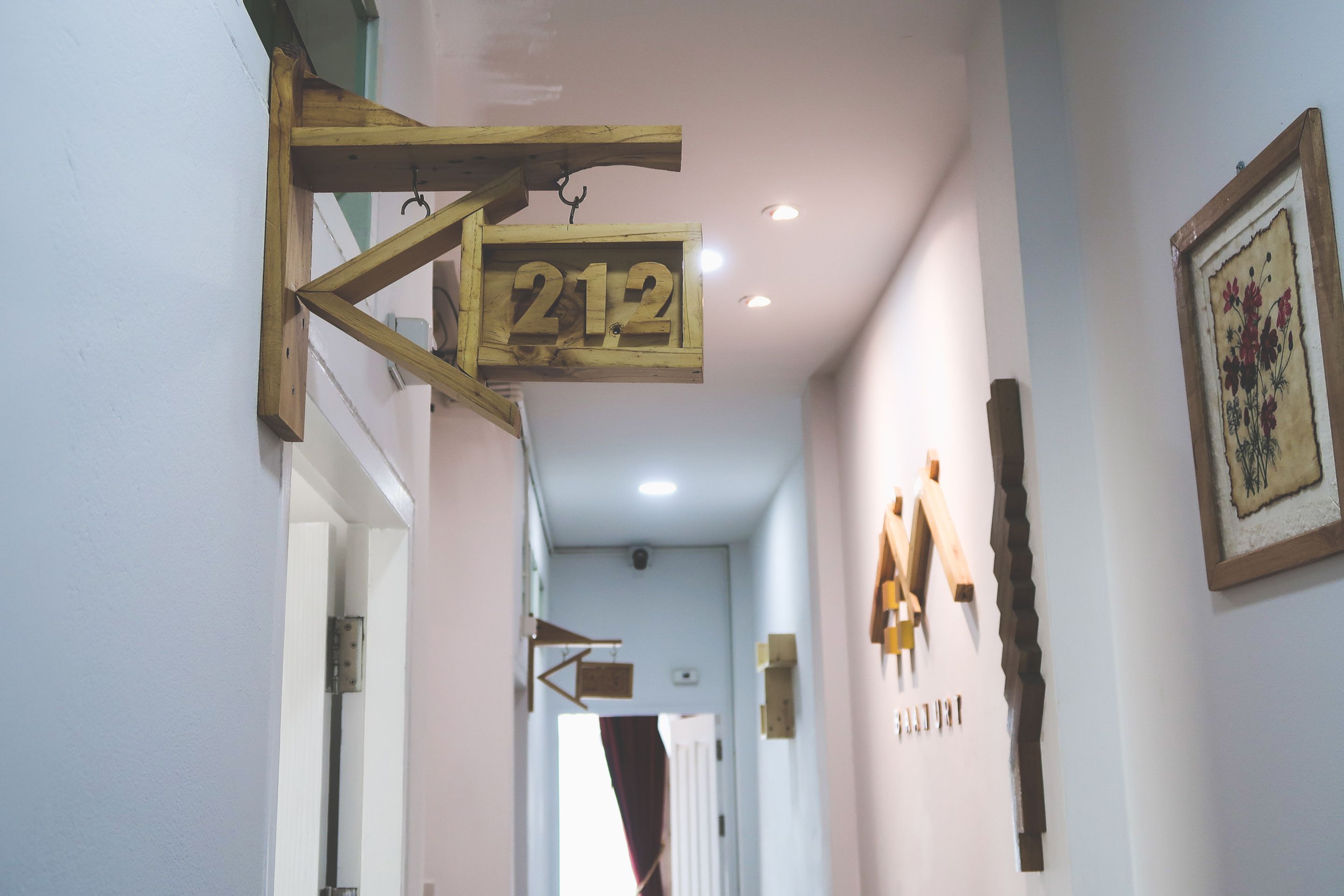 Um corredor com uma placa de madeira exibindo o número 212, uma obra de arte emoldurada na parede e uma porta ao fundo.
