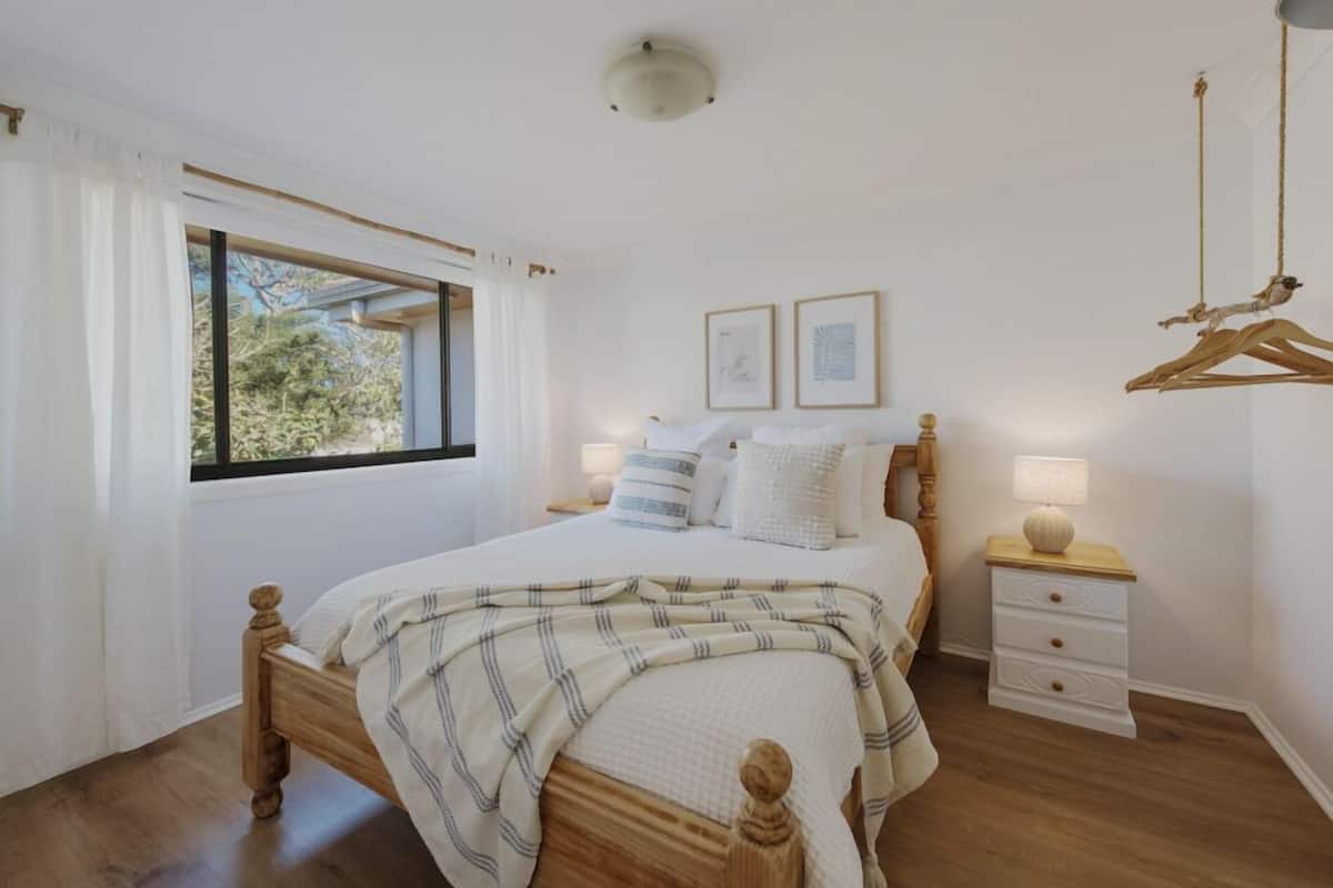 A bedroom with a wooden bed, white bedding, a bedside table with a lamp, and a window with curtains.