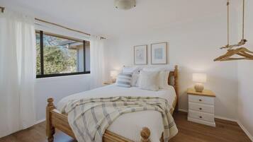 Un dormitorio con una cama de madera, ropa de cama blanca, una mesita de noche con lámpara y una ventana con cortinas.