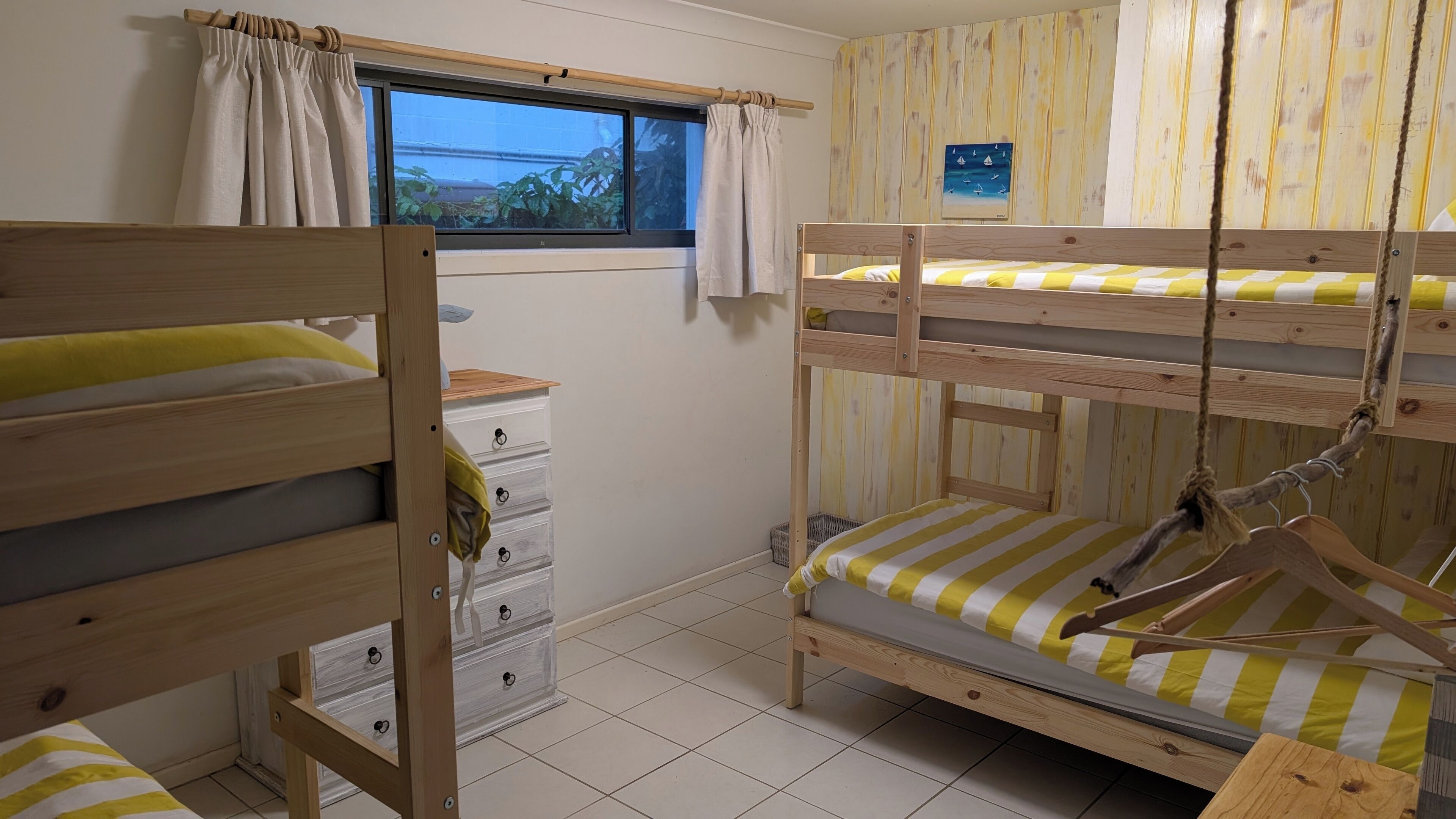 Een slaapkamer met stapelbed, een raam, een klein kastje en een aan de muur hangend plaatje.