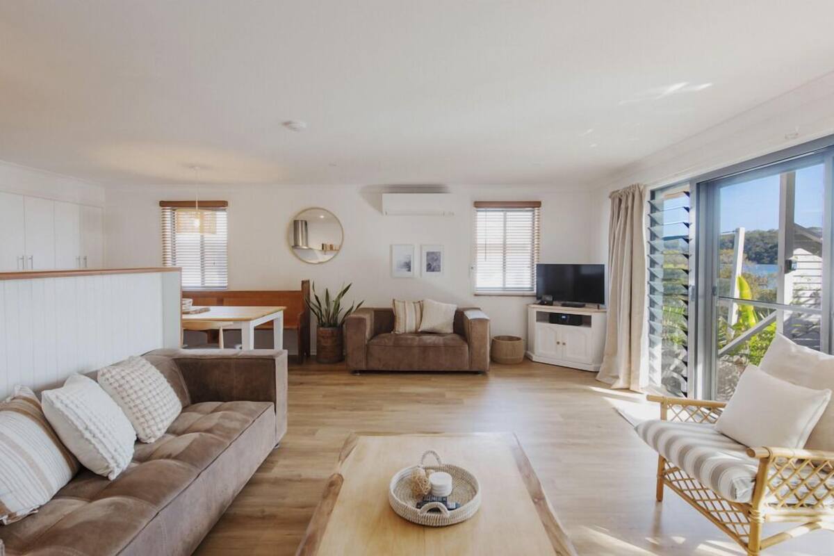 A modern living room with a sofa, a dining area, a TV, and a balcony with a view.
