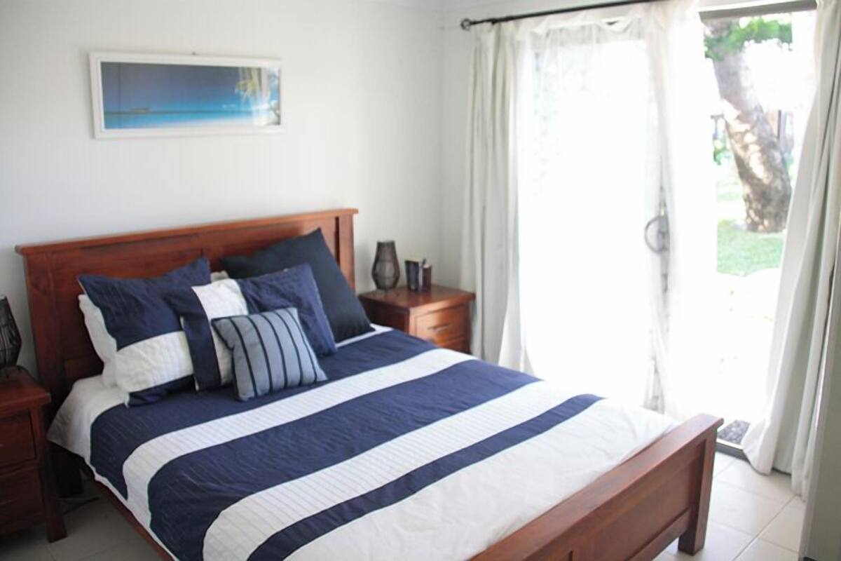 A neatly made bed with a striped bedspread and multiple pillows, a wooden headboard, a bedside table with a lamp, and a window with curtains.