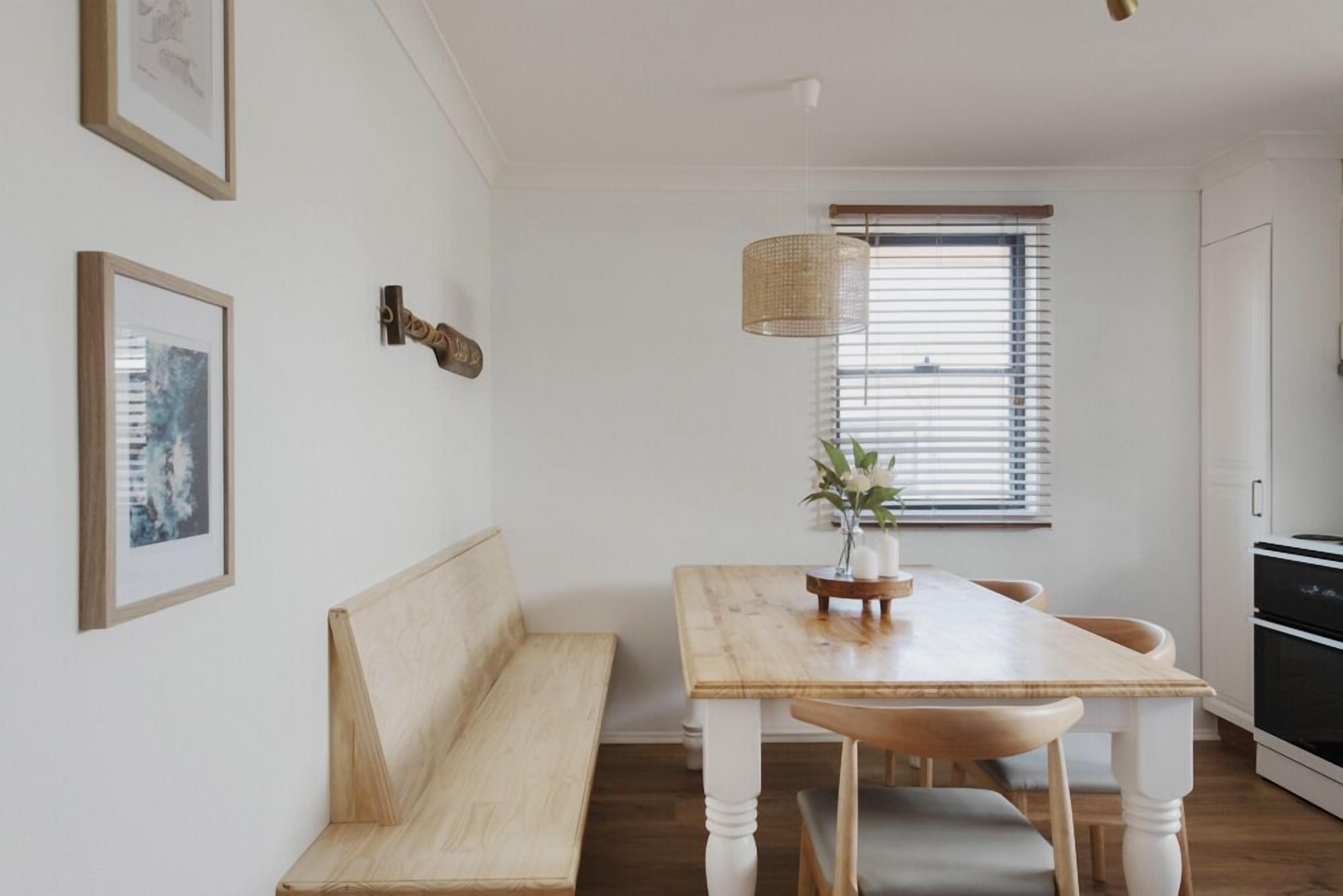 Een eethoek met een houten tafel en bank, een hanglamp en een raam met zonwering.