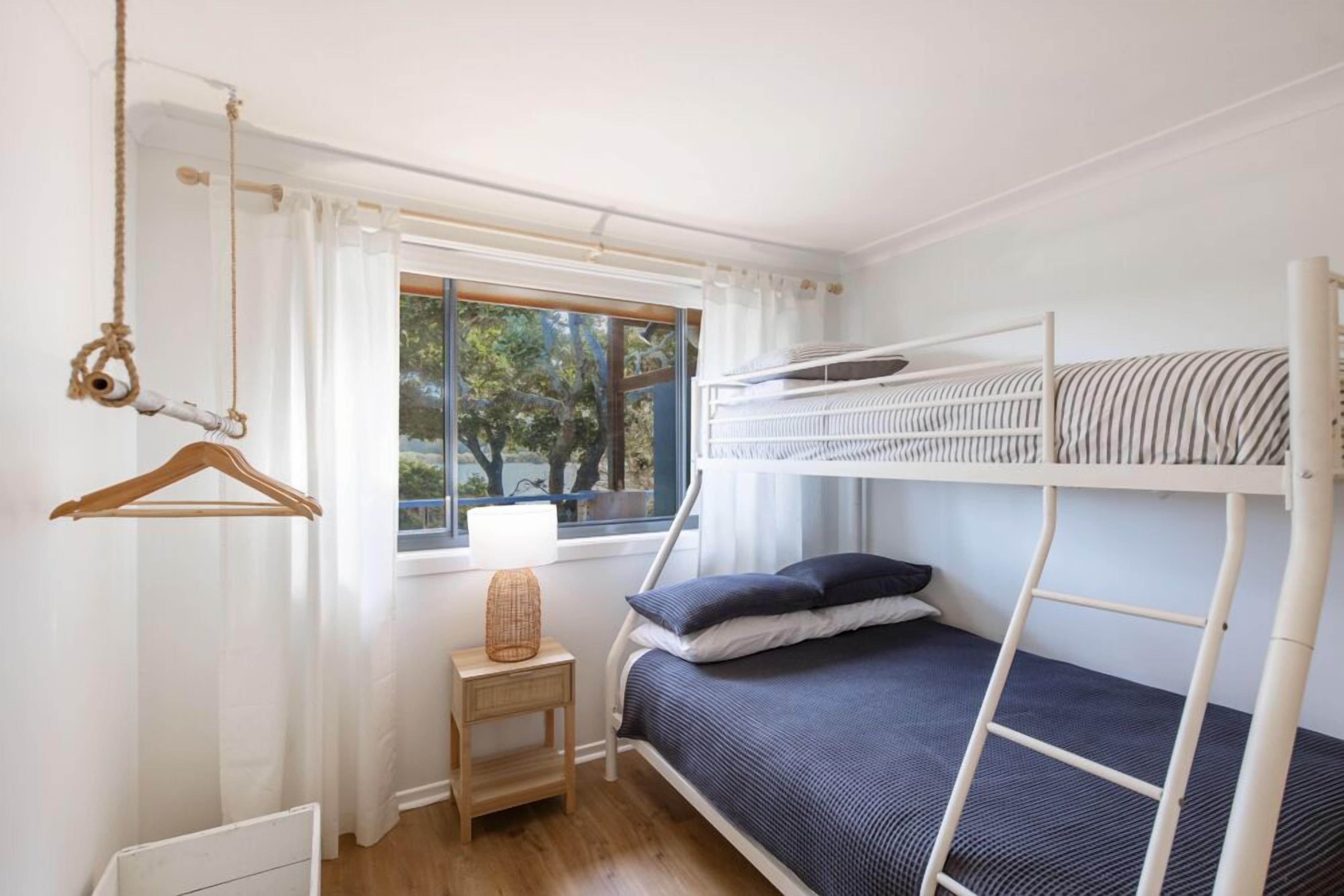 Habitación con literas, una ventana, una silla colgante de ratán, una mesita de noche con lámpara y suelo de madera.