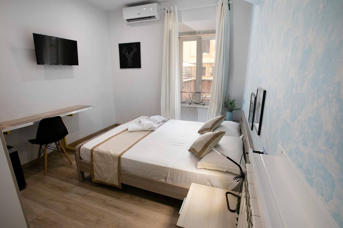 A hotel room with a bed, a desk, a chair, a TV, and a window with curtains.