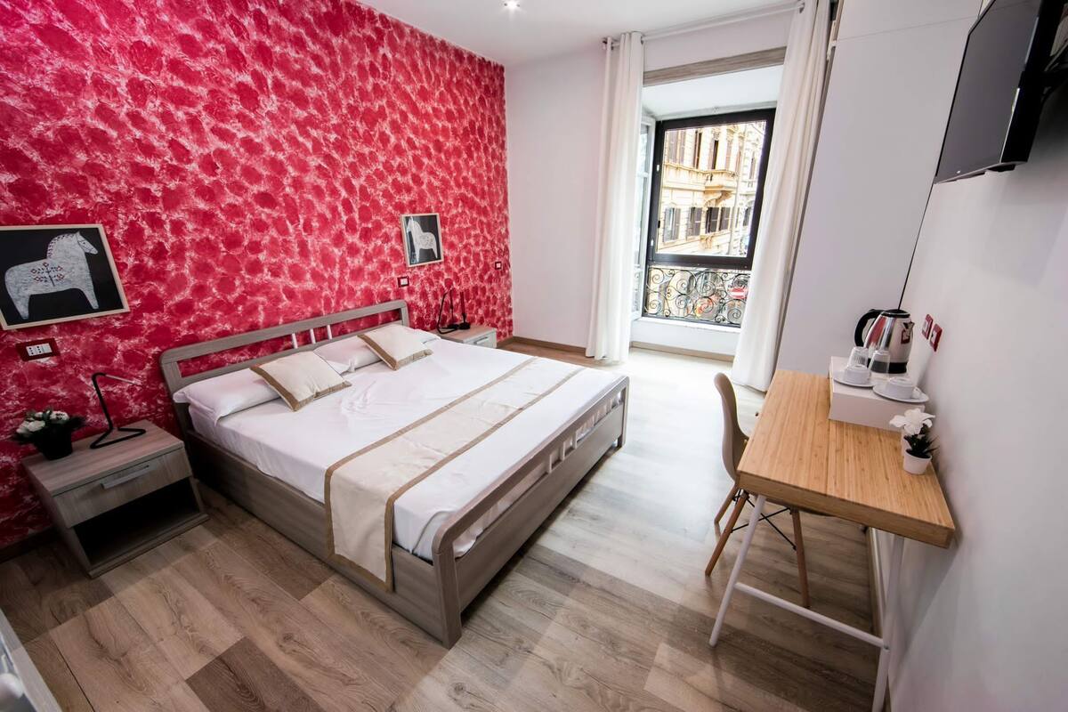 A bedroom with a bed, bedside tables, a desk, and a window with a view of buildings.