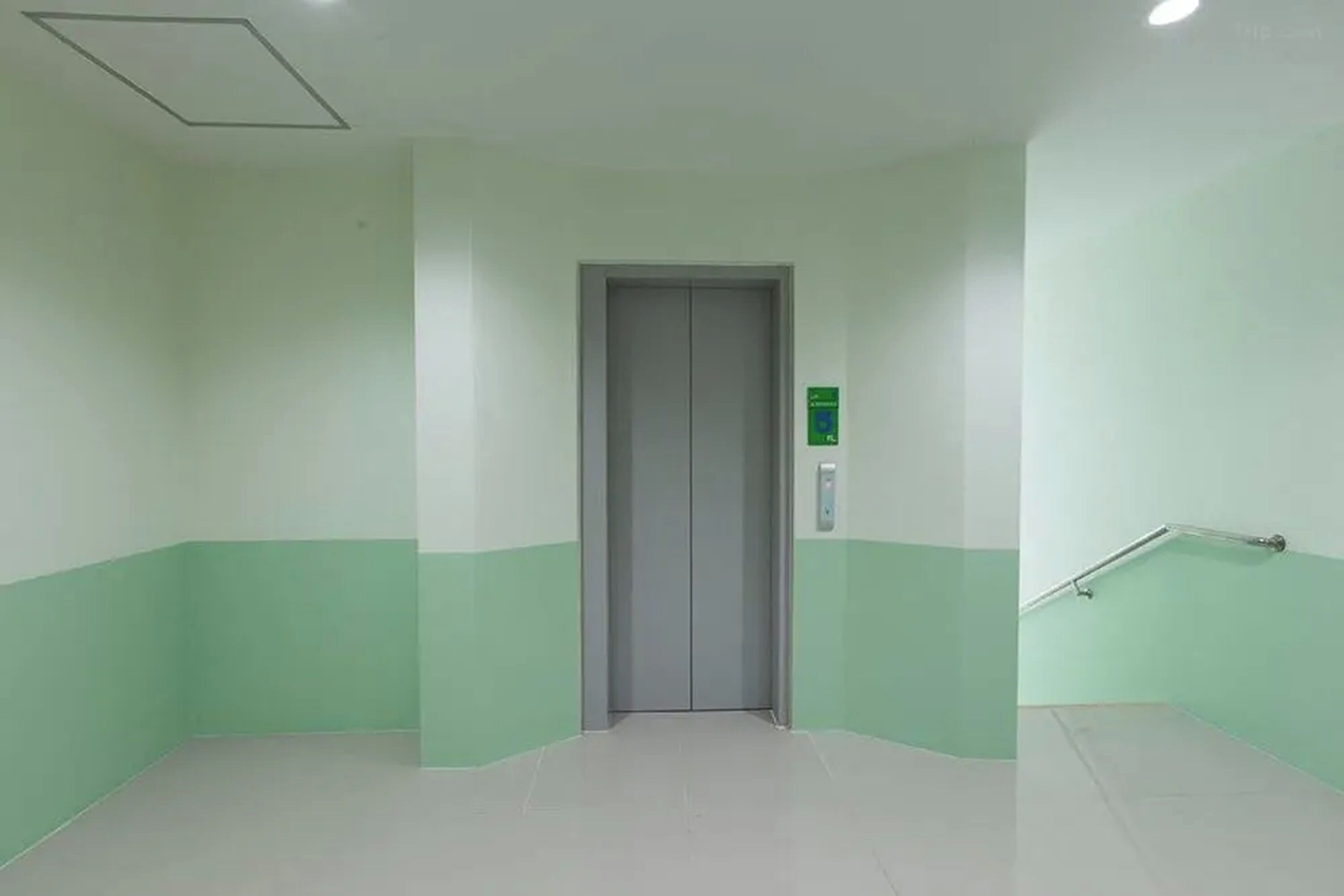 A hallway with a grey elevator, green walls, and a handrail on the right.