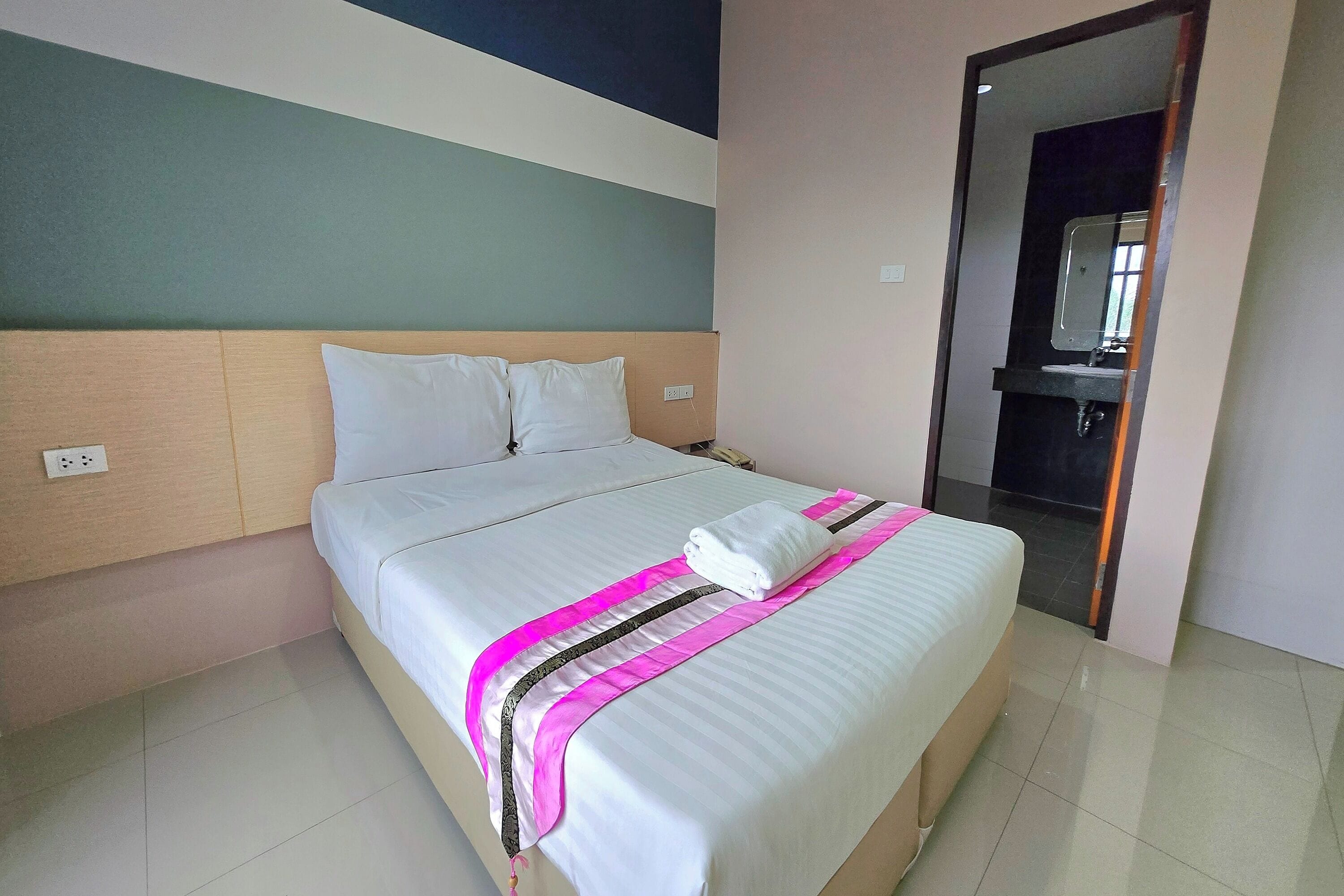 A neatly made bed with a white comforter and pink and black striped blanket, a wooden headboard, and a plain wall with a partial view of a bathroom mirror.