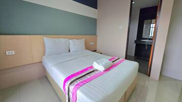 A neatly made bed with a white comforter and pink and black striped blanket, a wooden headboard, and a plain wall with a partial view of a bathroom mirror.
