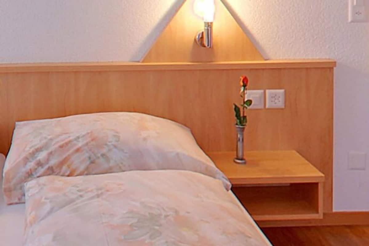 A bedroom with a wooden headboard, a bedside table with a vase of flowers, and a wall-mounted light fixture.