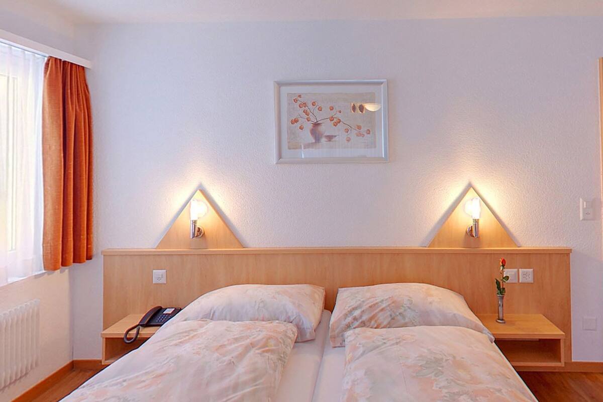 A double bed with two bedside tables, each with a lamp, in a room with a radiator, a phone, and a painting on the wall.