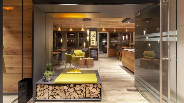 A modern interior with a central seating area featuring a fire feature, wooden logs, and a yellow bench. There are multiple seating options, including chairs and a sofa. The space includes a bar area with shelves and bottles. - Hotel Eden No. 7 (Saas-Fee)