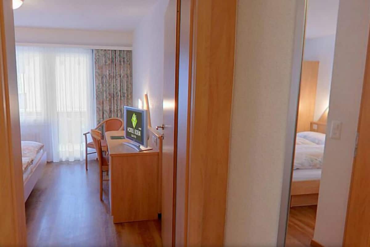 A hotel room with a bed, a desk with a computer, a chair, and a window with curtains.