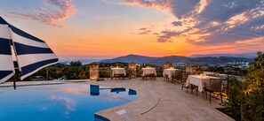 A poolside area with tables and chairs, overlooking a mountainous landscape at sunset.