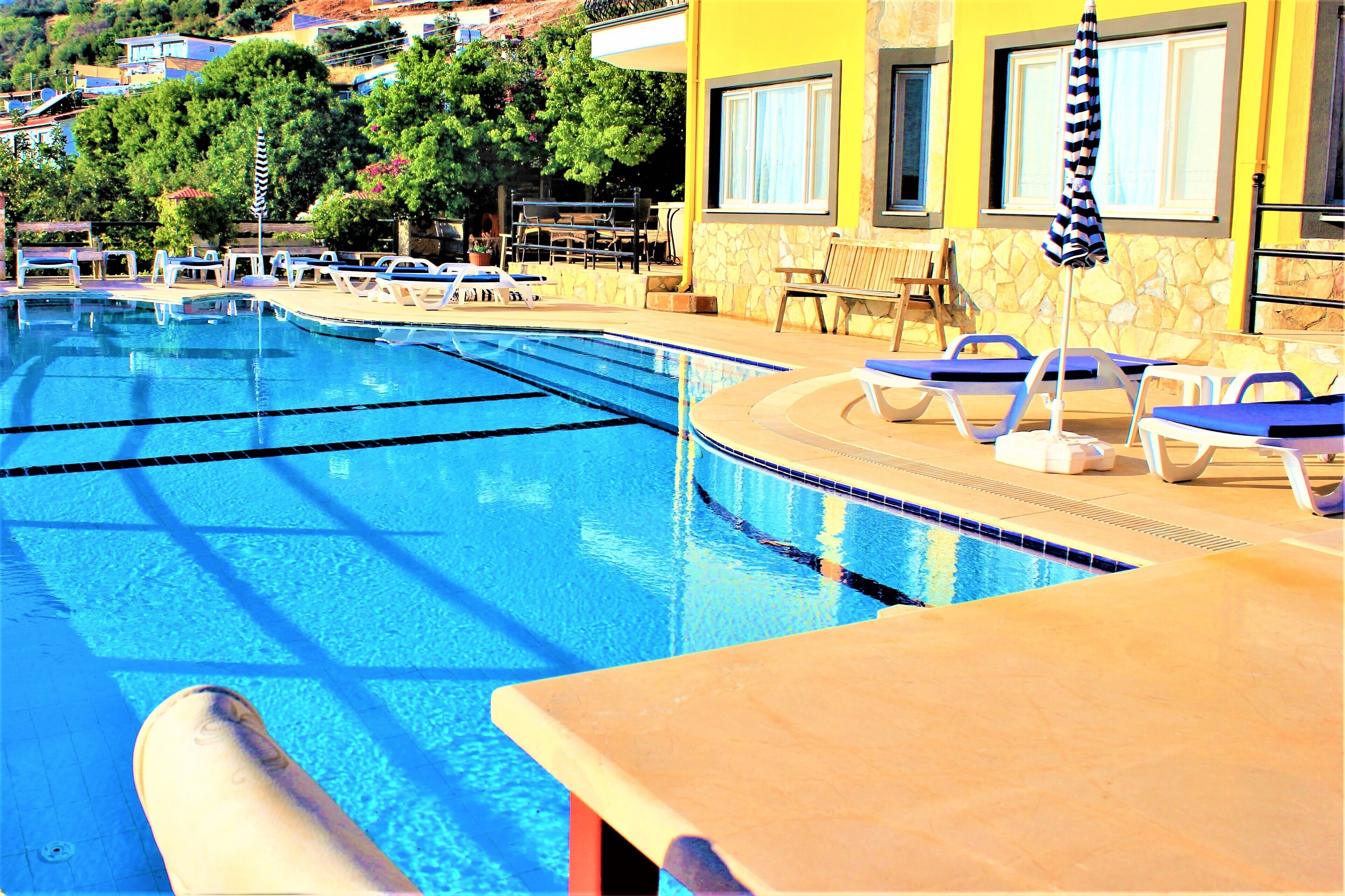 Una piscina con sillas de descanso y sombrillas, rodeada por un muro de piedra y un edificio amarillo.
