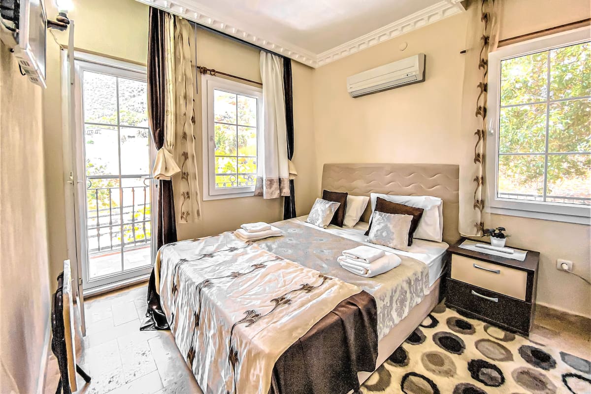A hotel room with a bed, a nightstand, an air conditioning unit, and a window with curtains.