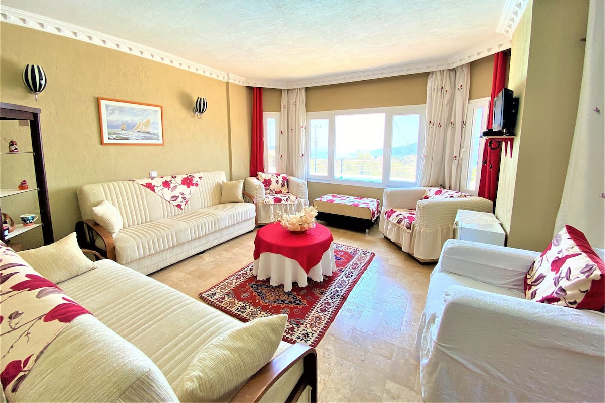 A well-lit living room with a sofa, armchairs, a coffee table, and a TV. There are windows with curtains, a painting on the wall, and a small shelf with decorative items.