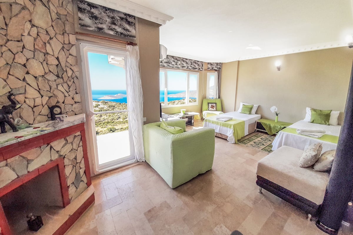 A hotel room with a stone wall, a balcony with a view of the sea, a bed with green bedding, a small table, and a chair.