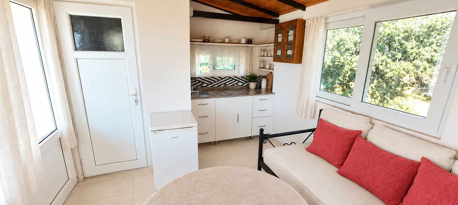 Habitación compacta con kitchenette, cama con almohadas rojas y ventana con vistas a zonas verdes.