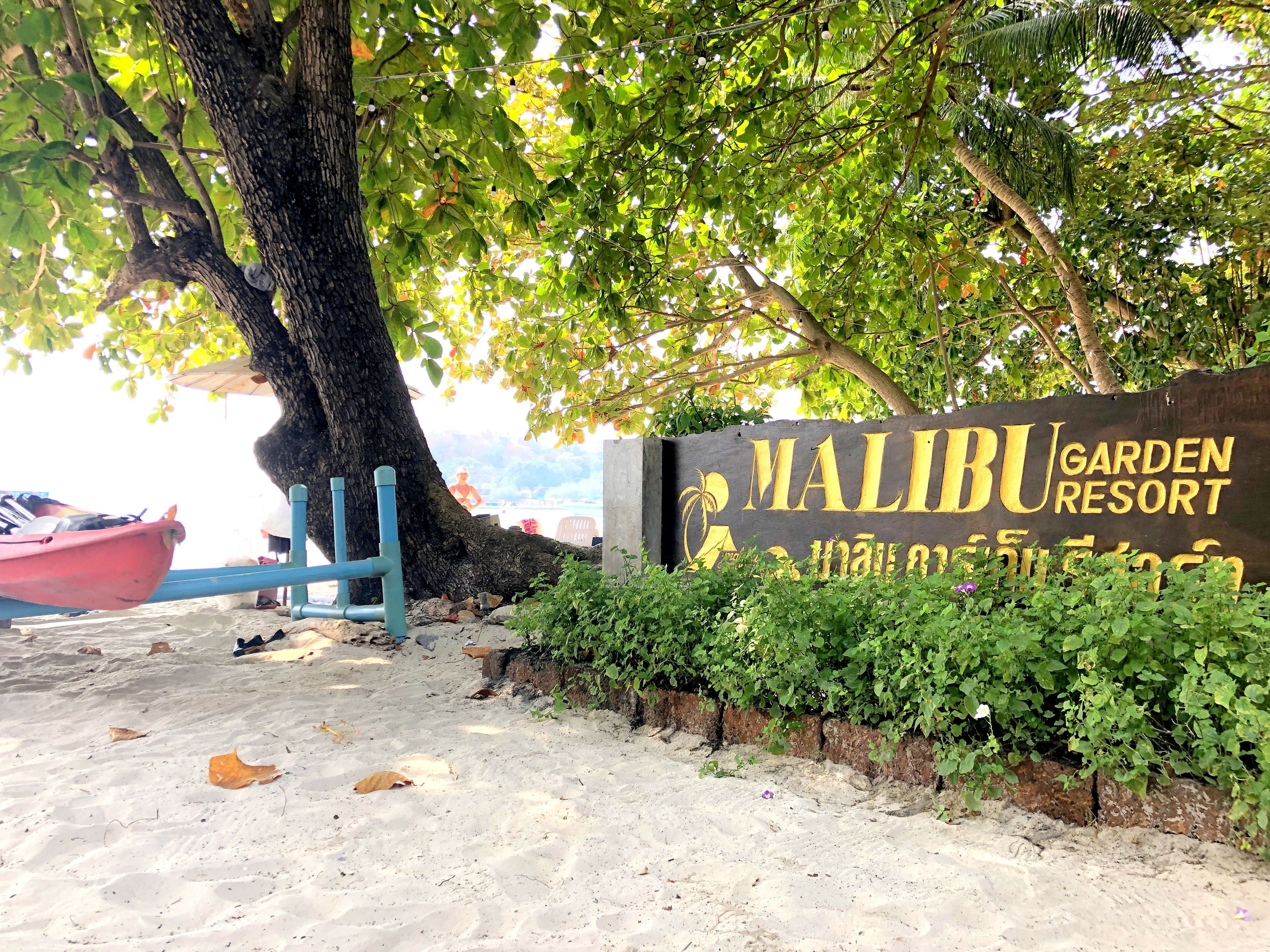 A sign for Malibu Garden Resort with a tree and a blue chair in the foreground.