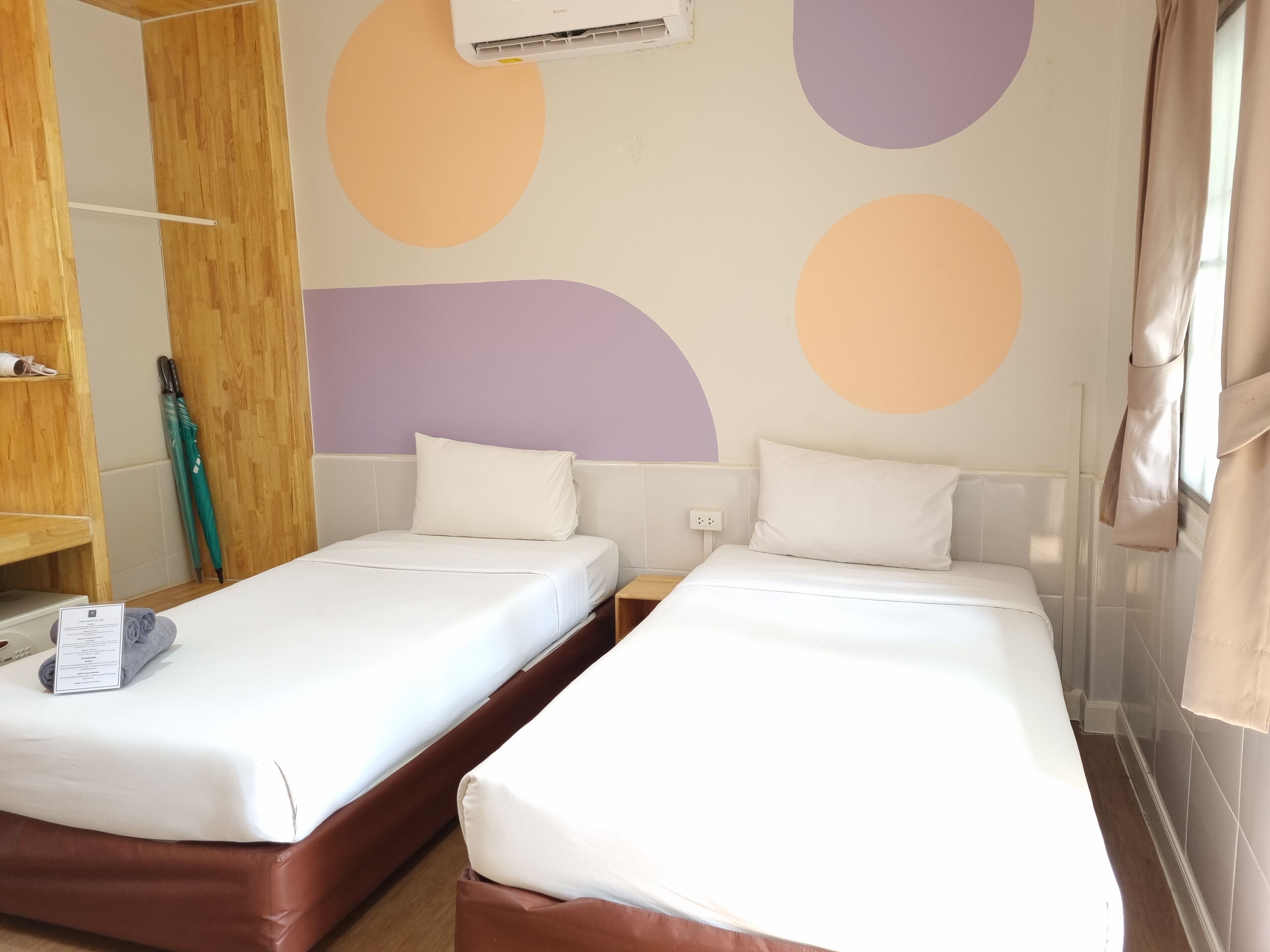 A room with two single beds, a wooden wardrobe, a green umbrella, and a wall with abstract shapes.