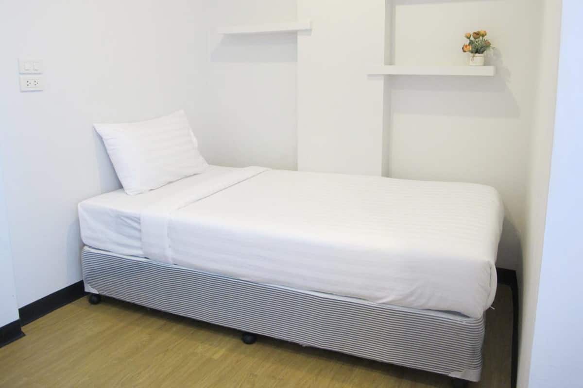 A single-bed room with a white bed, a small shelf with a flower, and a power outlet on the wall.