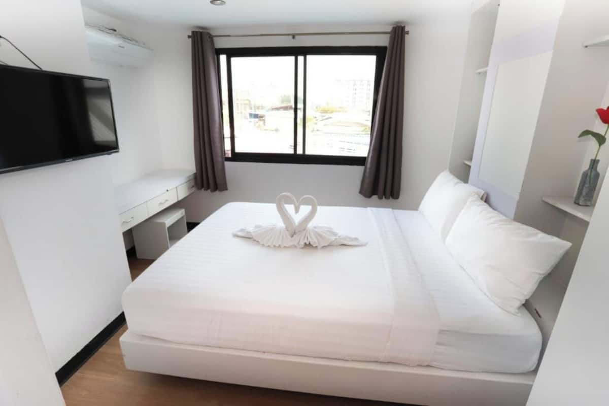 A neatly made bed with a heart-shaped decoration, a flat-screen TV on the wall, and a window with curtains.