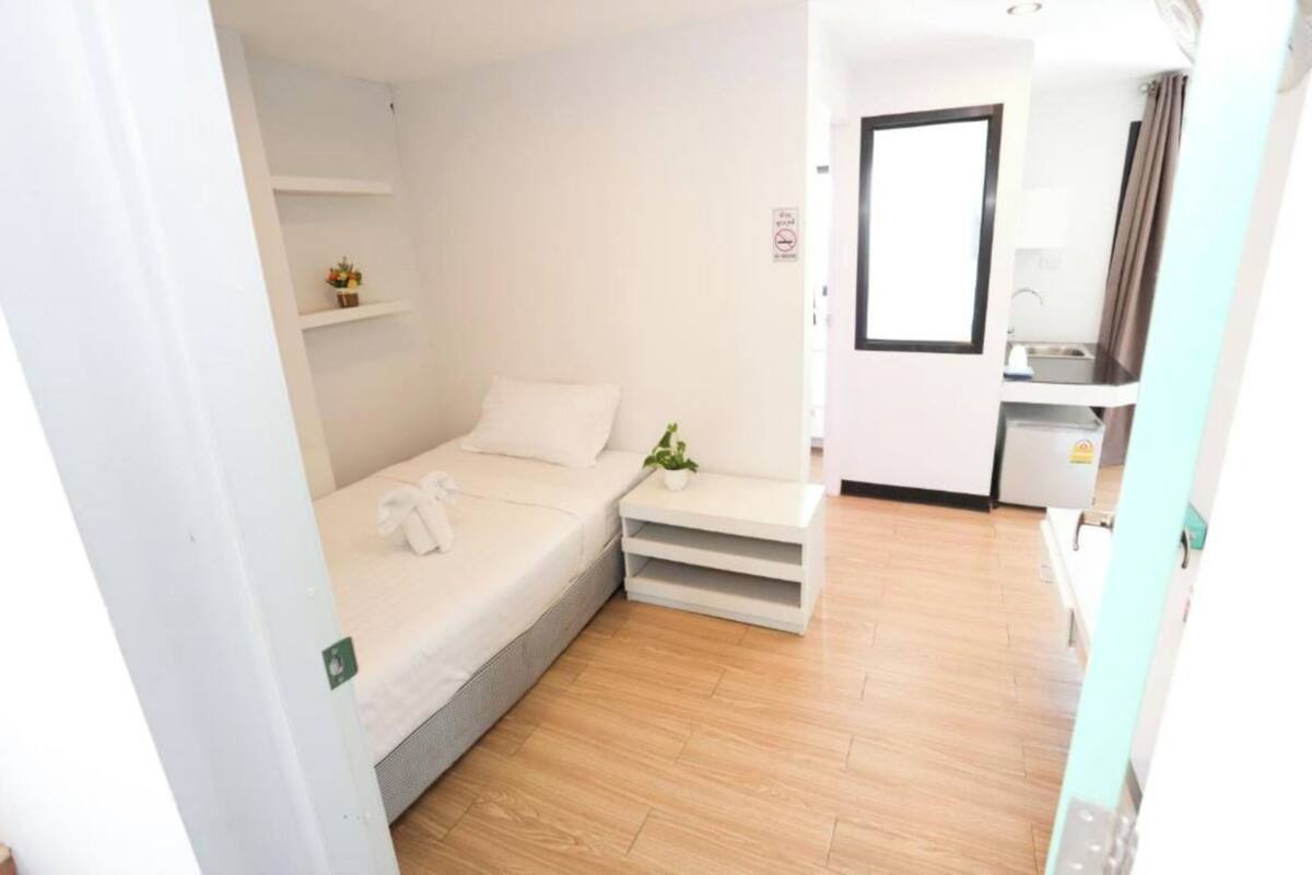 A compact hotel room with a bed, bedside table, a small plant, a mirror, and a kitchenette.
