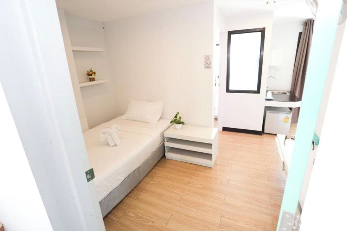 A compact hotel room with a bed, bedside table, a small plant, a mirror, and a kitchenette.