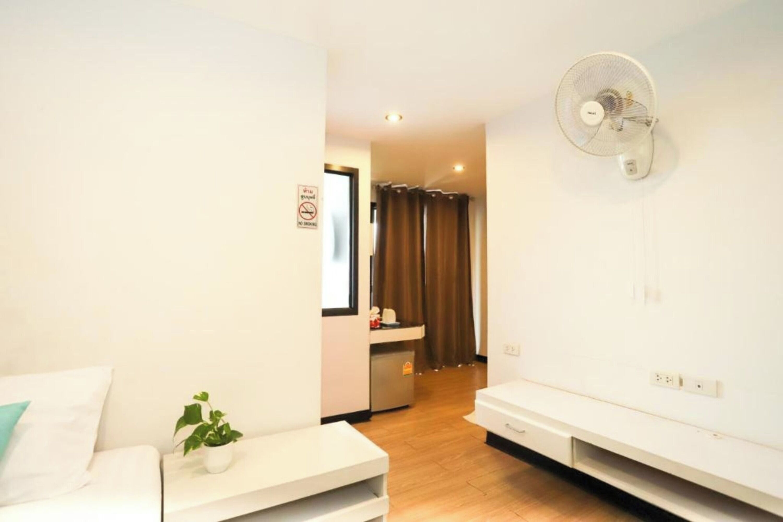 A hotel room with a bed, a small table with a plant, a wall-mounted fan, and a kitchenette in the background.