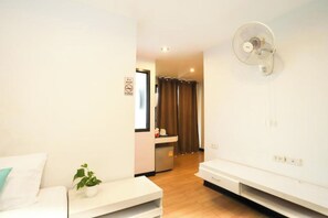 A hotel room with a bed, a small table with a plant, a wall-mounted fan, and a kitchenette in the background. - C Tower Hotel (Surat Thani)