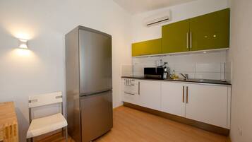 A compact kitchen with a stainless steel refrigerator, a microwave, and a sink. There is a wooden chair and a small table. The cabinets are white with one green door.