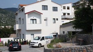 Un edificio de varios pisos con fachada blanca y tejas rojas. Un vehículo blanco está estacionado frente al edificio, con el logotipo 