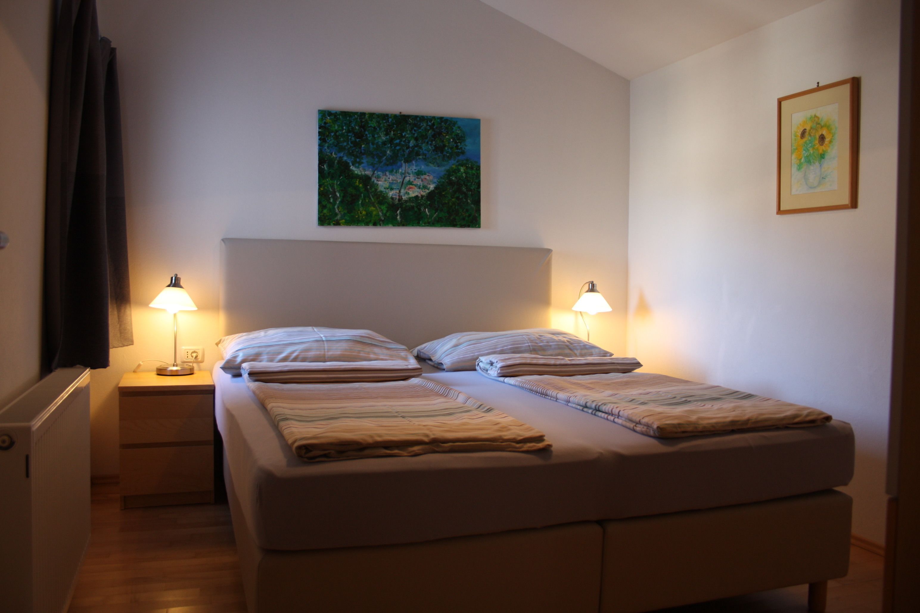 A double bed with two bedside tables, each with a lamp, a painting above the bed, and a framed picture on the wall.