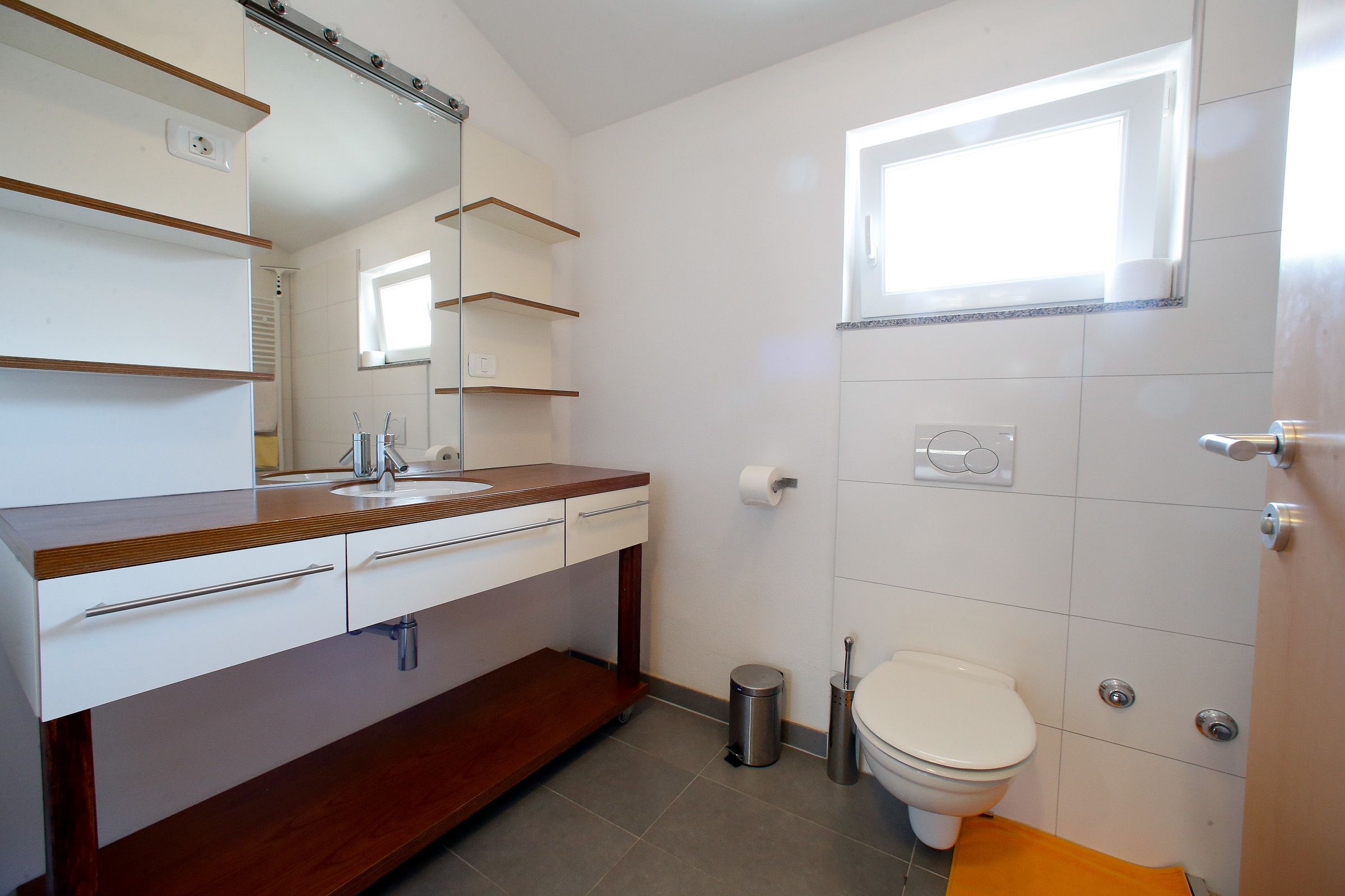 A bathroom with a wooden vanity, a toilet, a trash can, and a window.