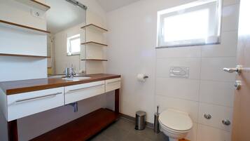 A bathroom with a wooden vanity, a toilet, a trash can, and a window.