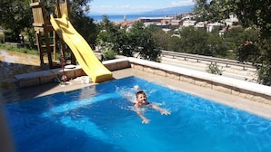 Una piscina con un tobogán amarillo, rodeada por una solana de hormigón y una vista de un pueblo y montañas al fondo.
